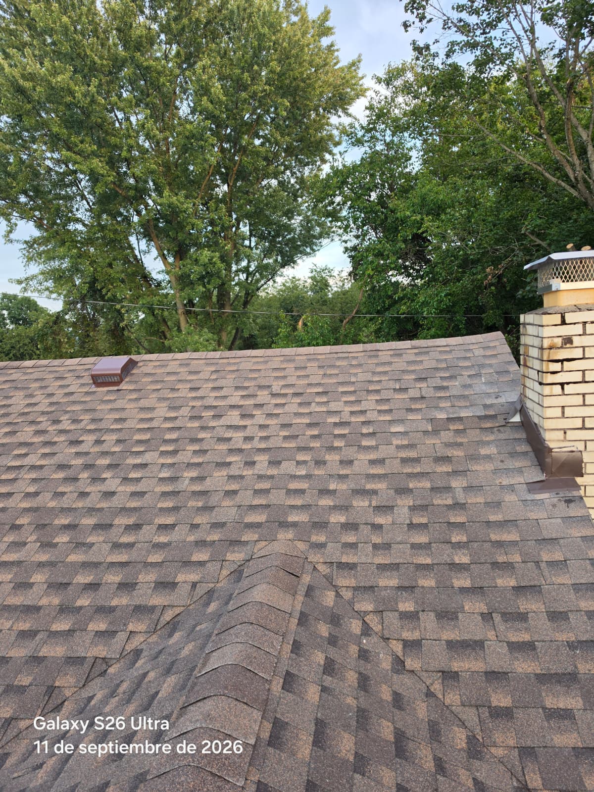 Hipped roof intersection with brown architectural shingles, ridge caps and a brick chimney with mesh cap