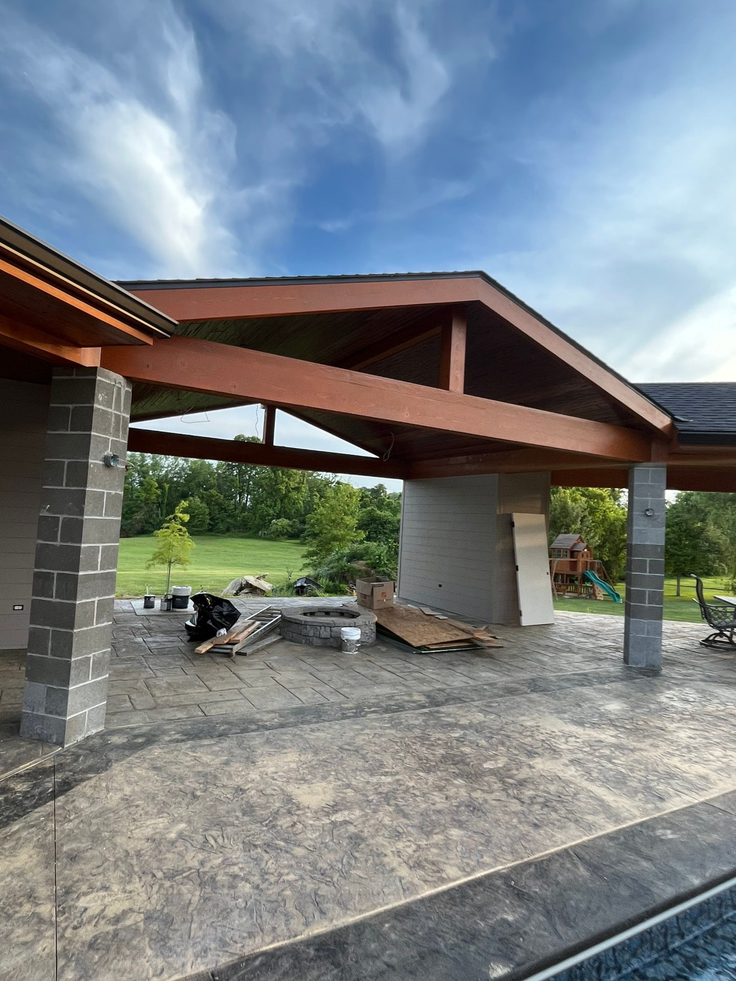 Heavy timber pavilion framing with stained beams and metal roof over a stamped concrete patio